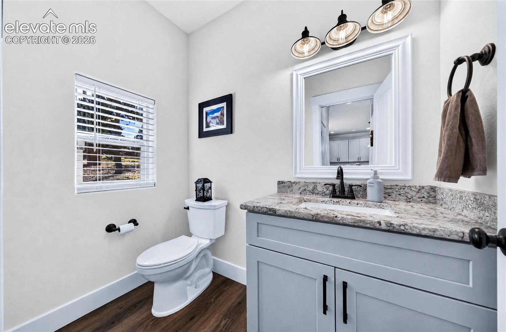 Vanity featuring a stone-look countertop, rectangular undermount sink, and matte black hardware, paired with wood-finish flooring, a three-light fixture, and a framed mirror