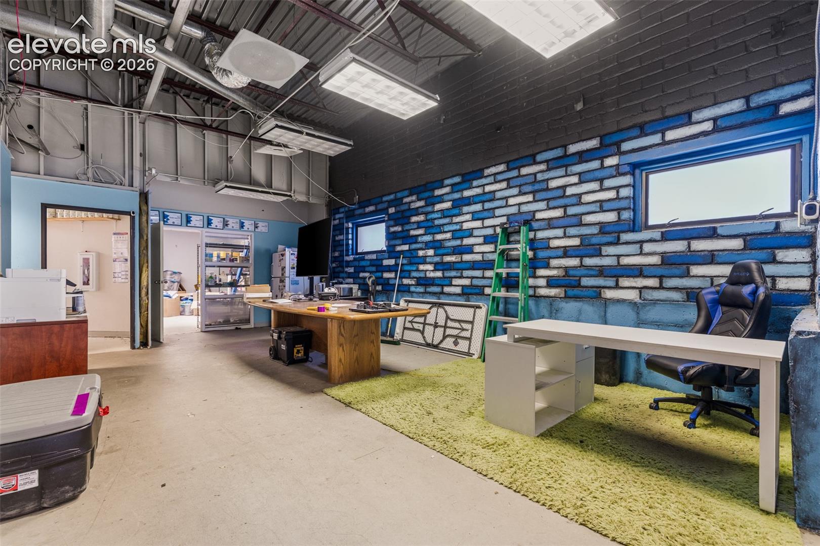 Exposed ceiling structure with industrial ductwork, painted brick masonry accent wall, high-output industrial lighting fixtures, and concrete flooring