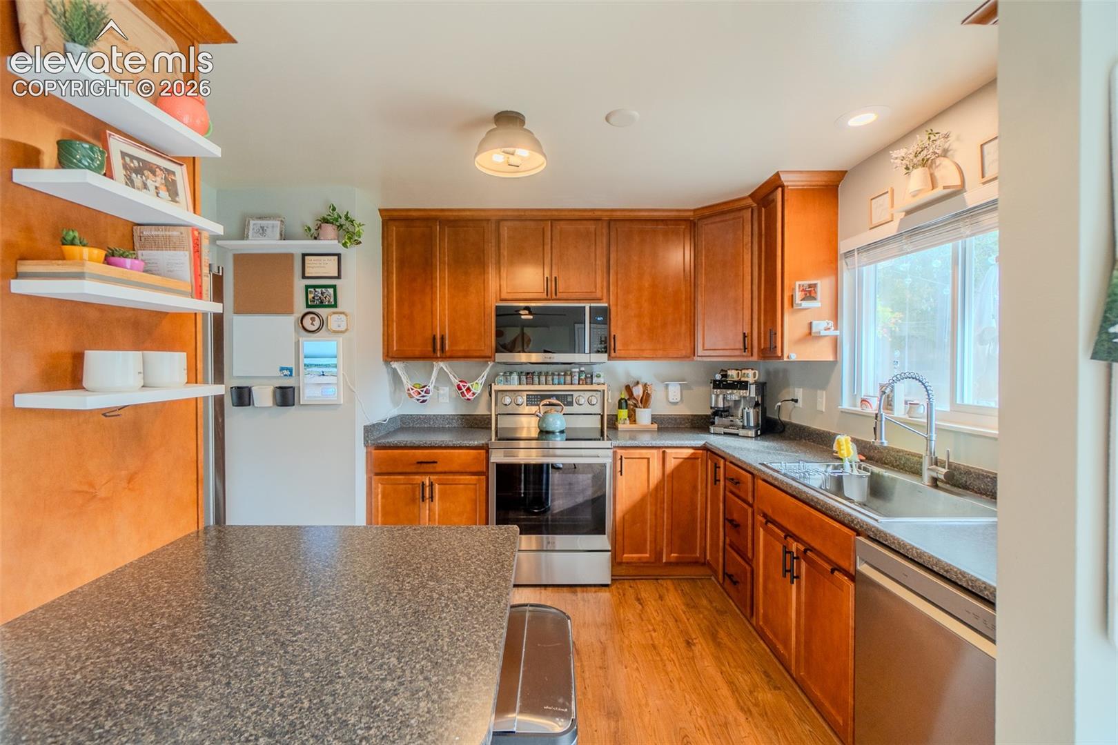 Kitchen featuring wood cabinetry, stainless steel appliances, speckled stone-finish countertops, floating wall shelving, and wood-finish flooring