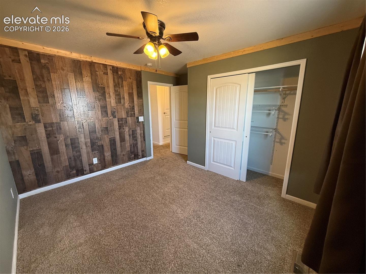 Accent wall featuring vertical wood-finish planking, carpeted flooring, ceiling fan with integrated lighting, and closet with sliding paneled doors and built-in wire shelving