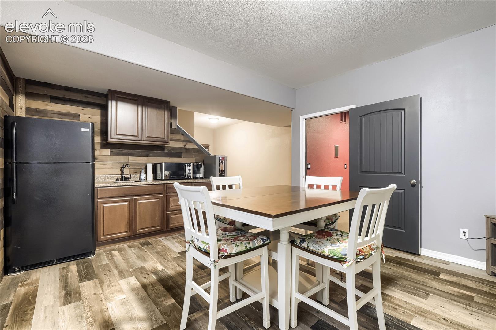 Lower-level wet bar featuring wood-look paneling, a stainless steel sink, wood-finish flooring, dark wood cabinetry, and a granite countertop