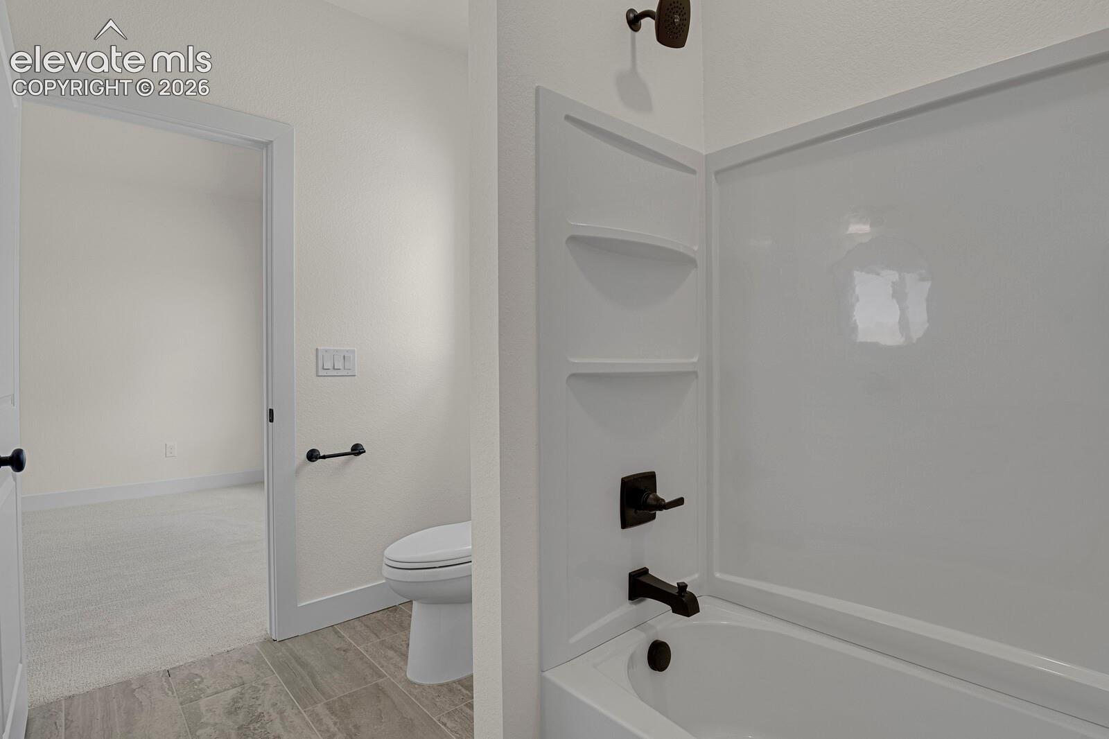 Bathroom featuring a tub-shower combination with matte black fixtures, integrated shelving, an elongated toilet, and neutral-tone tile flooring