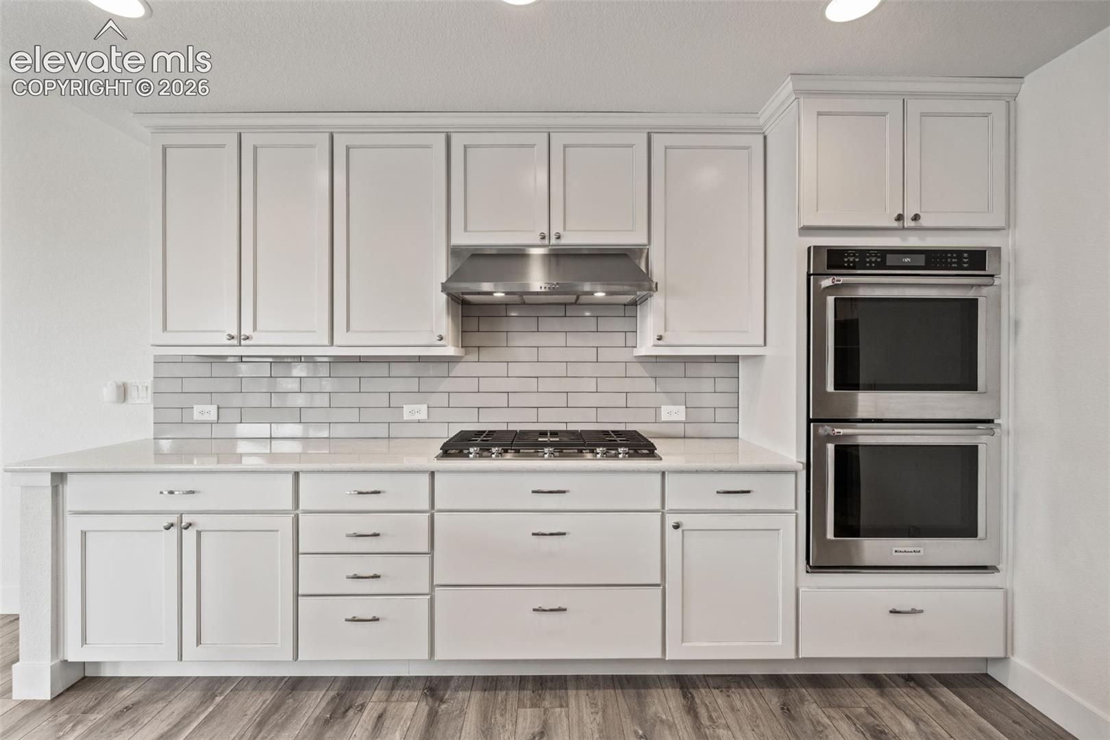 White shaker cabinetry, light subway tile backsplash, stainless steel gas cooktop, stainless steel range hood, and double wall ovens