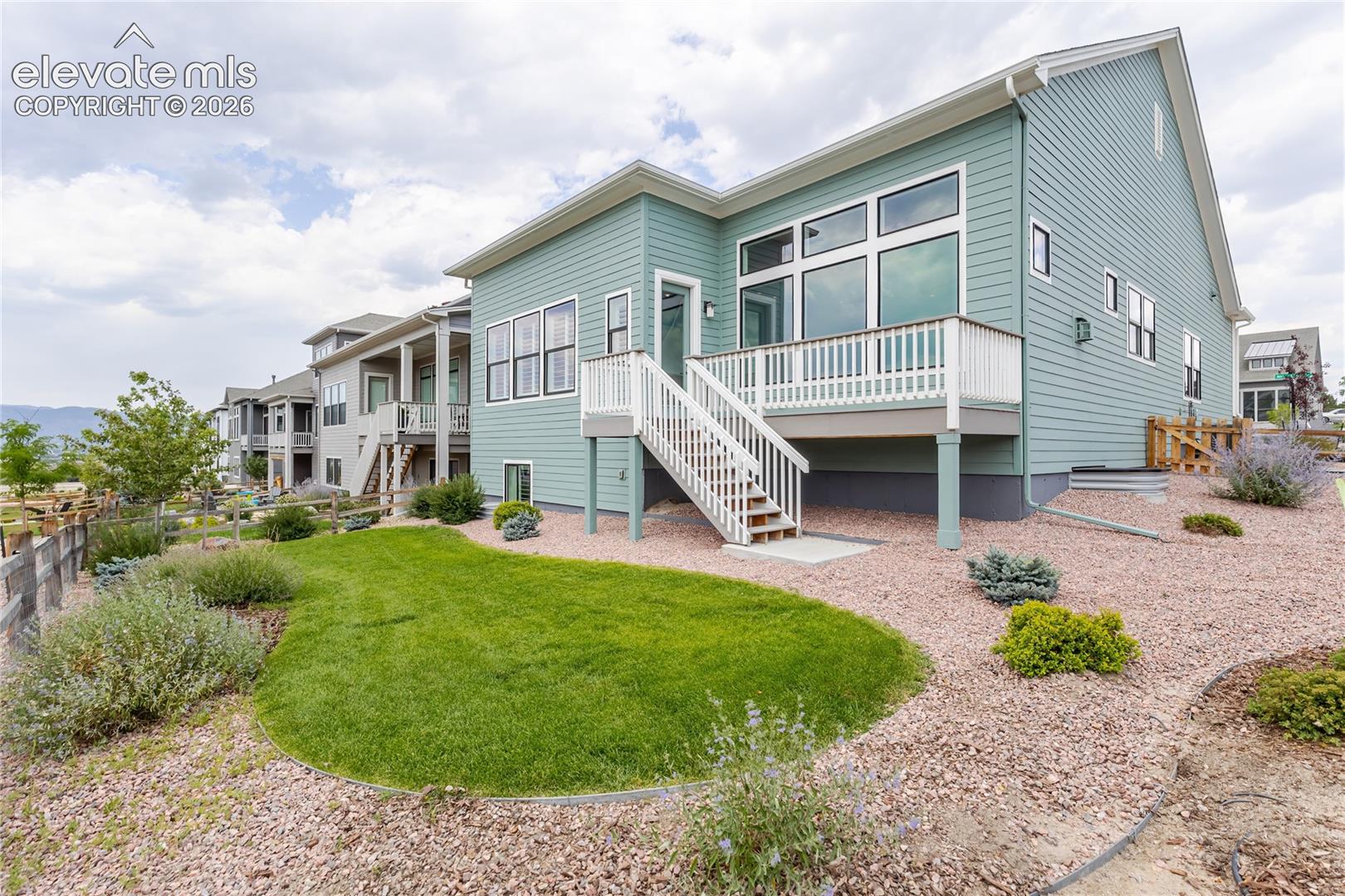 Rear exterior featuring a spacious deck with white railings and stairs, extensive large-pane windows, light green siding, and a landscaped yard with a manicured lawn and gravel beds