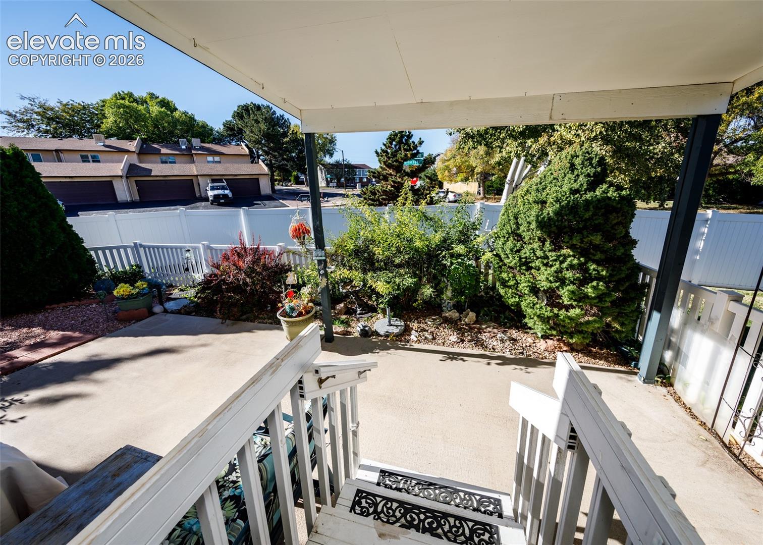 Covered concrete patio featuring white vinyl fencing, mature landscaping, and structural support posts