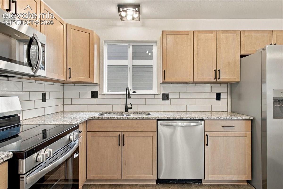 Kitchen featuring light wood-finish cabinetry, stone-pattern countertops, a white subway tile backsplash, stainless steel appliances, and a matte black faucet