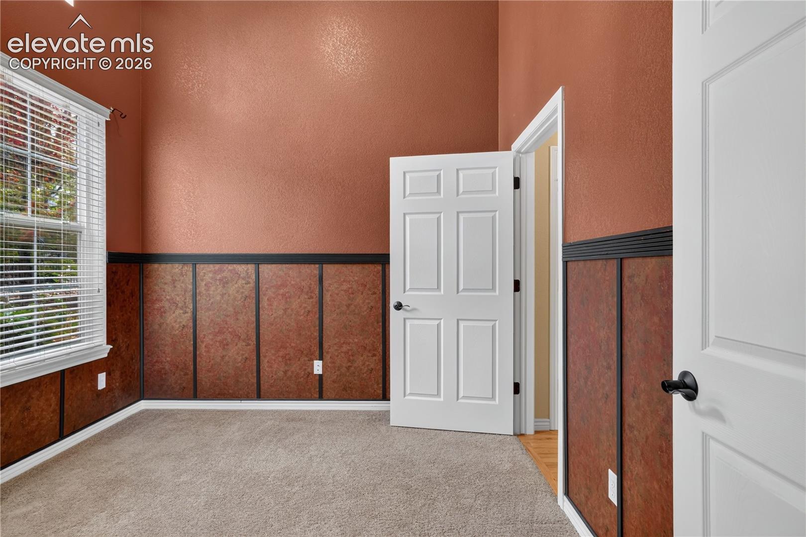 Office - featuring high ceilings, textured wall paneling with chair rail, neutral carpeting, and a multi-pane window, with attached half bath