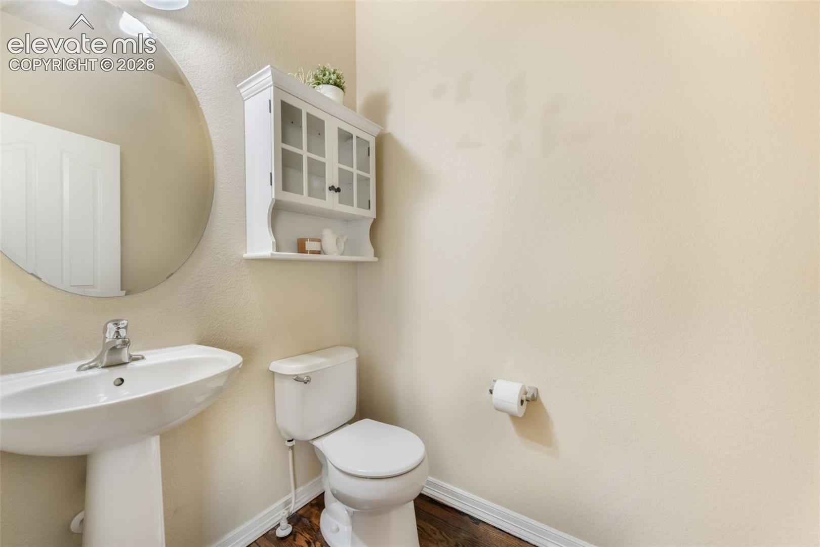 Pedestal sink with chrome faucet, circular mirror, wall-mounted storage cabinet with glass-front doors, and wood-finish flooring