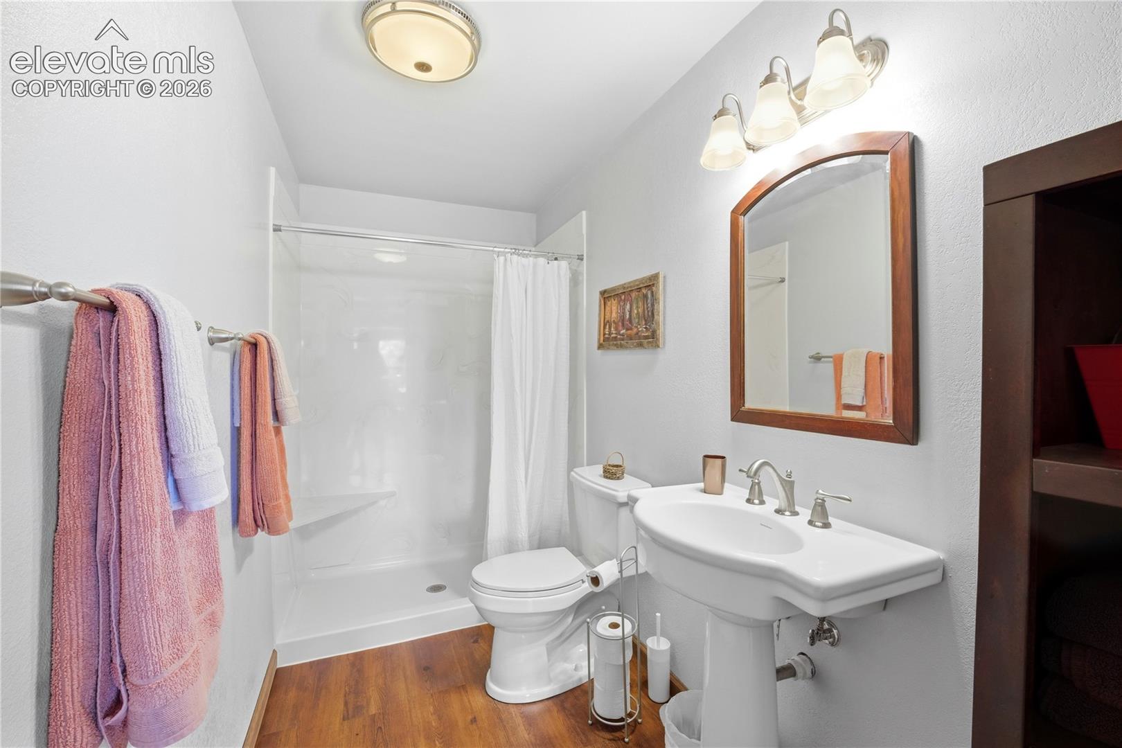 Pedestal sink with chrome fixtures, wood-finish flooring, white shower with clear enclosure, wall-mounted light fixture, and recessed shelving