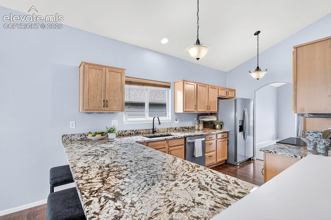 Kitchen featuring natural wood-finish cabinetry, stone-patterned countertops, a stainless steel refrigerator, matte black faucet hardware, and dual pendant light fixtures