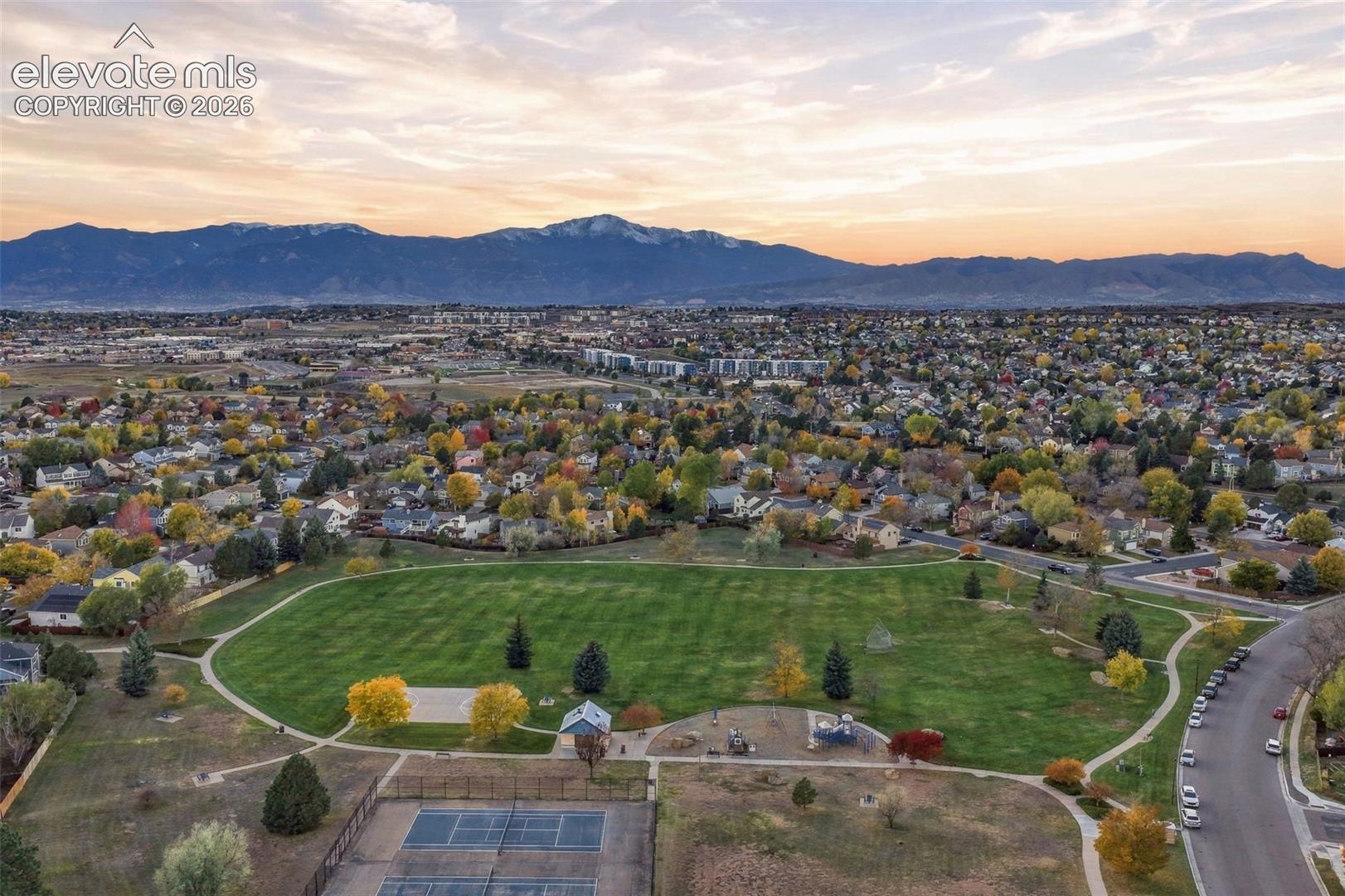 Aerial perspective of a neighborhood park featuring expansive green space, paved walking paths, a hard-surface tennis court, and a mountain range backdrop