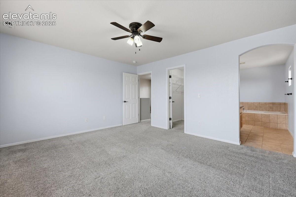 Spacious interior with neutral carpeting, a ceiling fan, a walk-in closet, and a tiled soaking tub alcove
