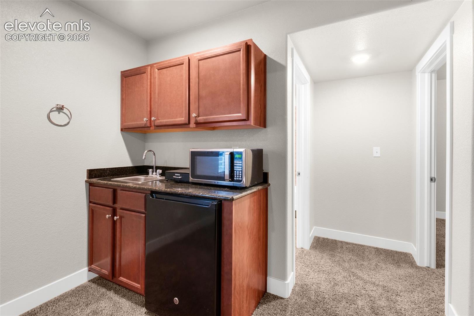 Kitchenette featuring dark-stained wood cabinetry, stone-finish countertops, stainless steel sink with metal faucet, and carpeted flooring