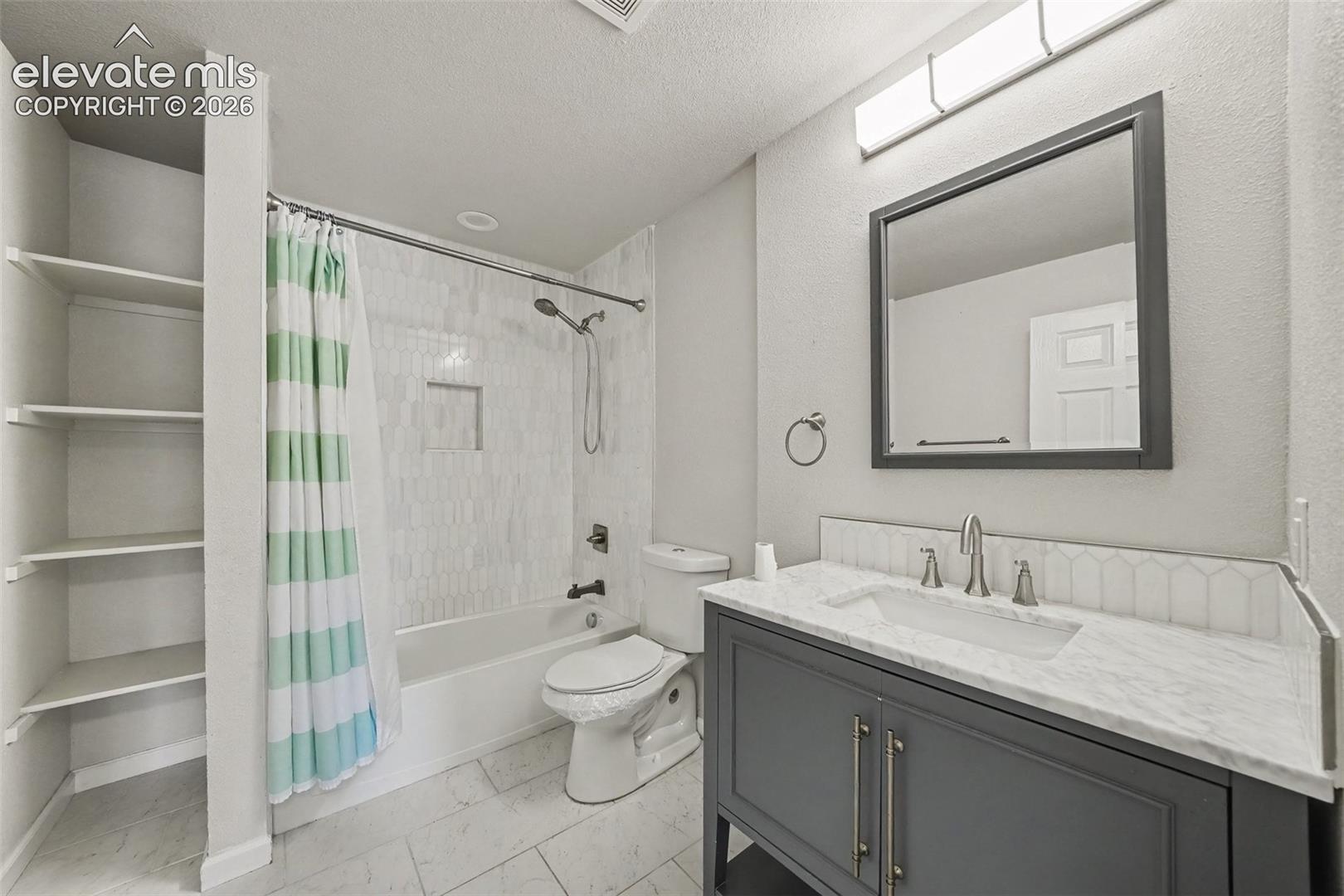 Bathroom featuring a vanity with stone countertop, rectangular undermount sink, built-in shelving, tiled shower surround with recessed niche, and tile flooring