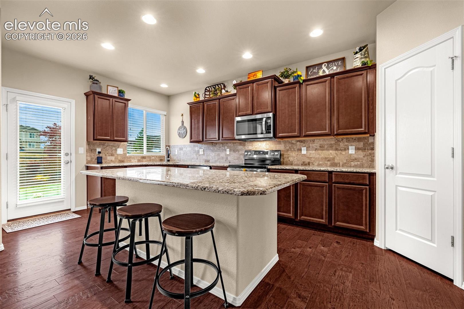 Kitchen featuring dark wood cabinetry, a central island with a stone-finish countertop, stone-tile backsplash, stainless steel appliances, and wood-finish flooring
