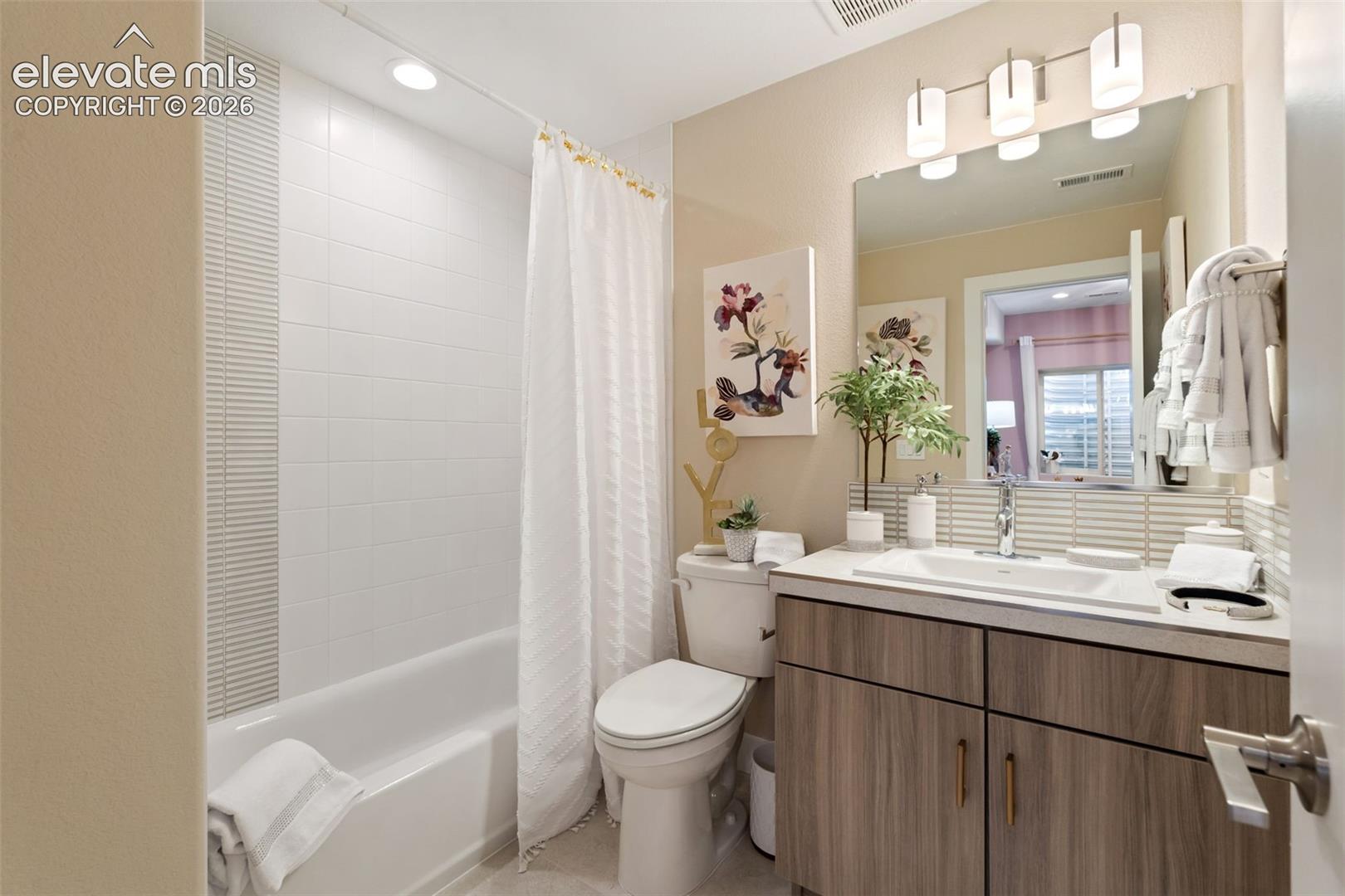 Bathroom featuring a vanity with wood-finish cabinetry, a white integrated countertop sink, a full-width wall mirror, multi-light fixture, and a tiled shower surround