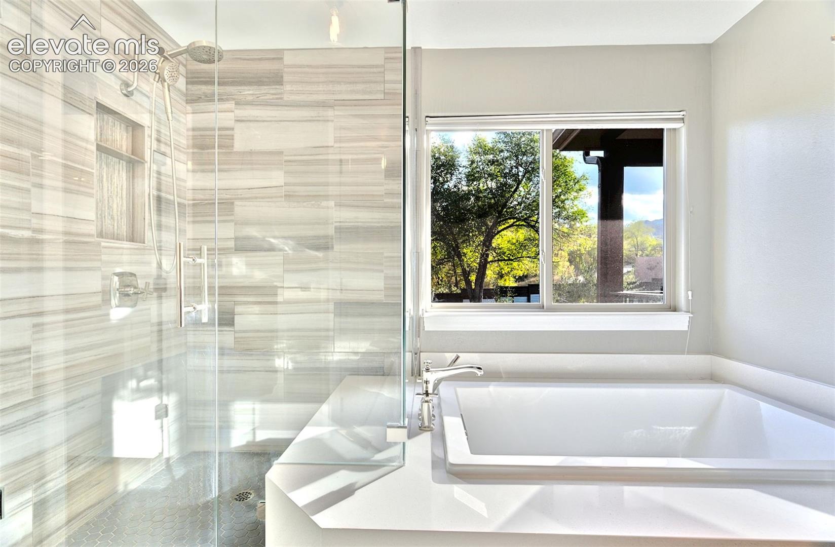 Walk-in glass-enclosed shower with horizontal tile surround and integrated wall niche, accompanied by a drop-in soaking tub with a solid-surface deck and polished chrome fixtures