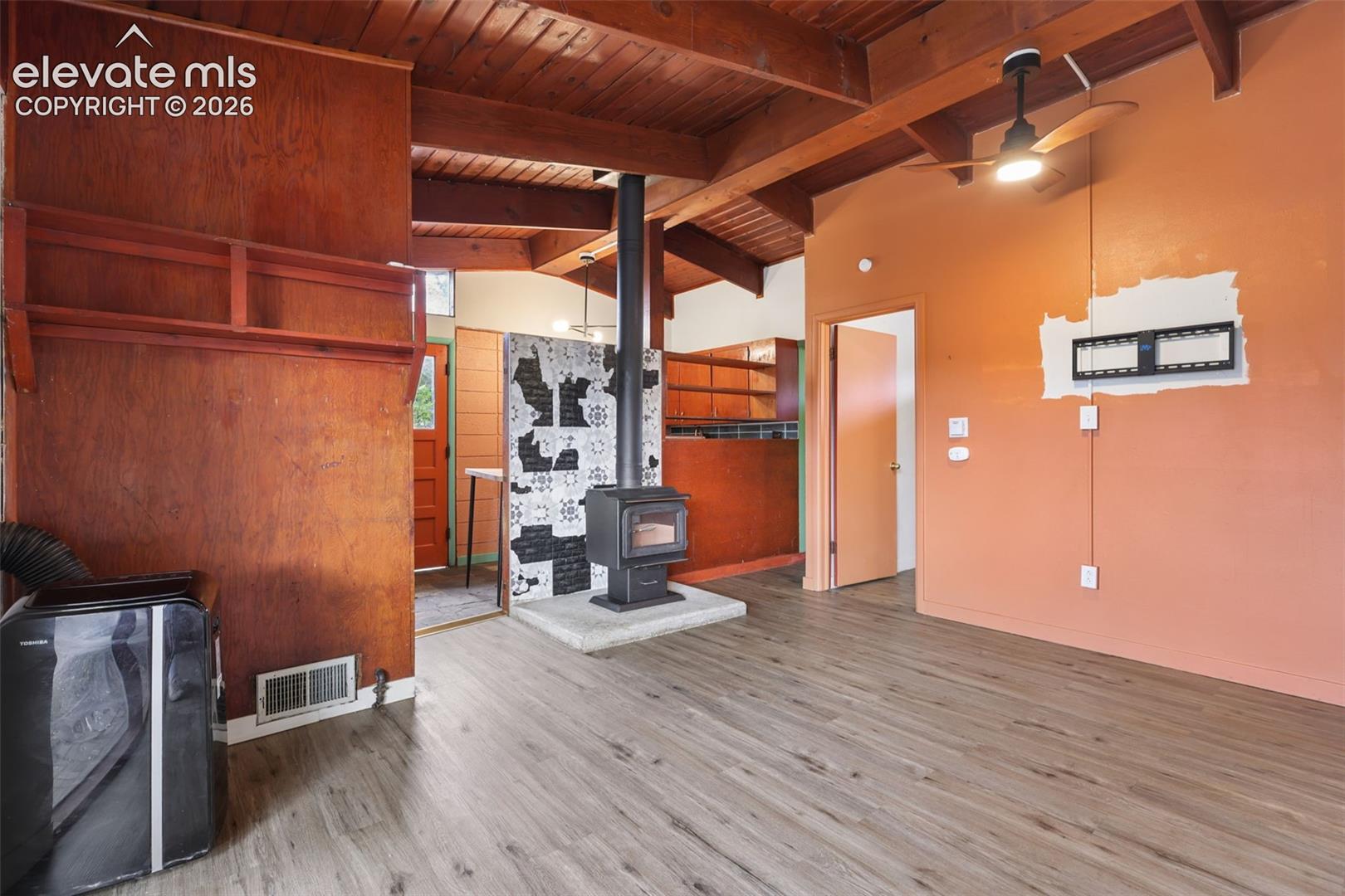 Wood-finish flooring, exposed wood beam ceiling, contemporary ceiling fan with light, and a freestanding wood-burning stove