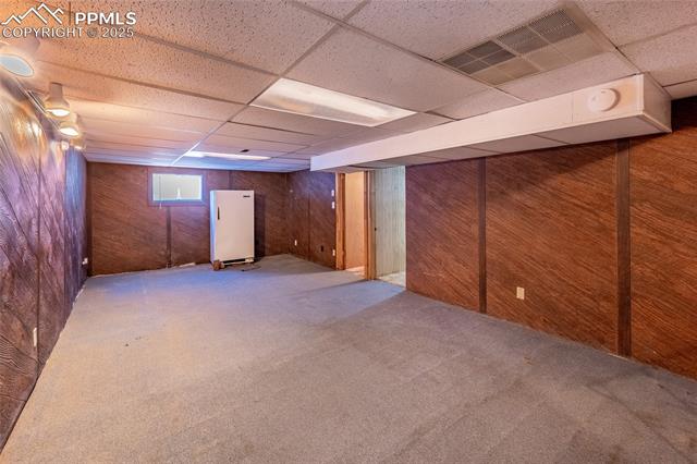 Below grade area with wooden walls, a paneled ceiling, carpet floors, and freestanding refrigerator