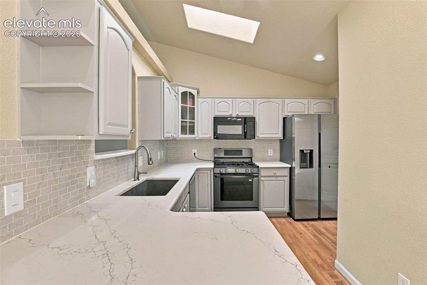 Kitchen featuring quartz countertops, white cabinetry with glass-front detailing, a subway tile backsplash, a vaulted ceiling with a skylight, and wood-finish flooring