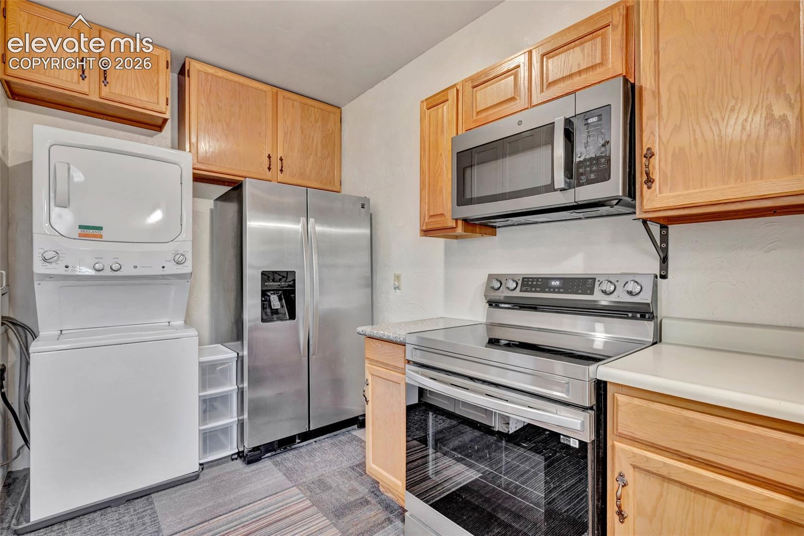 Kitchen featuring light-finish wood cabinetry, stainless steel appliances, a stacked laundry unit, and stone-finish countertops