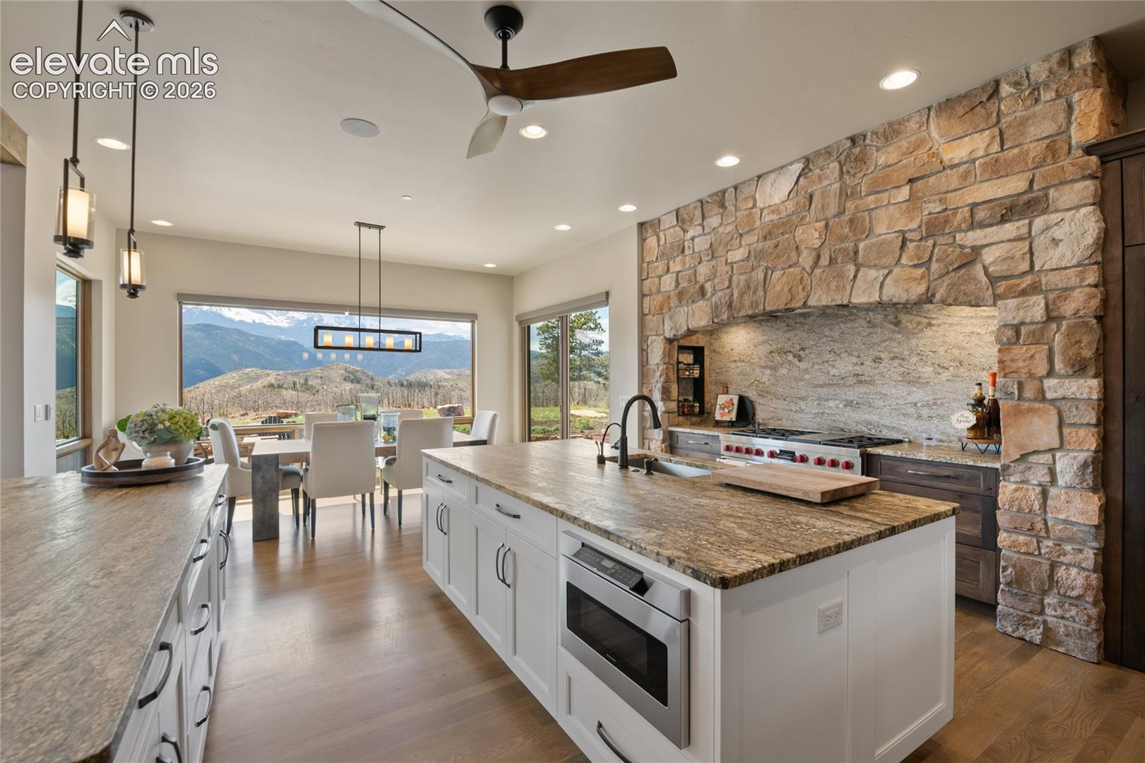 Open-concept kitchen and dining with Wolf range, full-slab granite backsplash, stone accent wall, and panoramic Pikes Peak views from the dining area.
