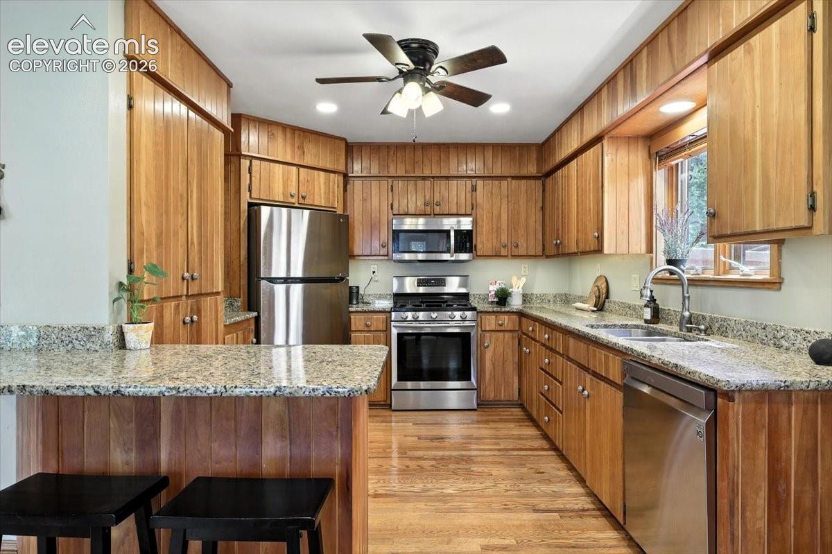 Kitchen featuring natural wood cabinetry, granite countertops, stainless steel appliances, wood-finish flooring, and a ceiling-mounted light fixture with integrated fan