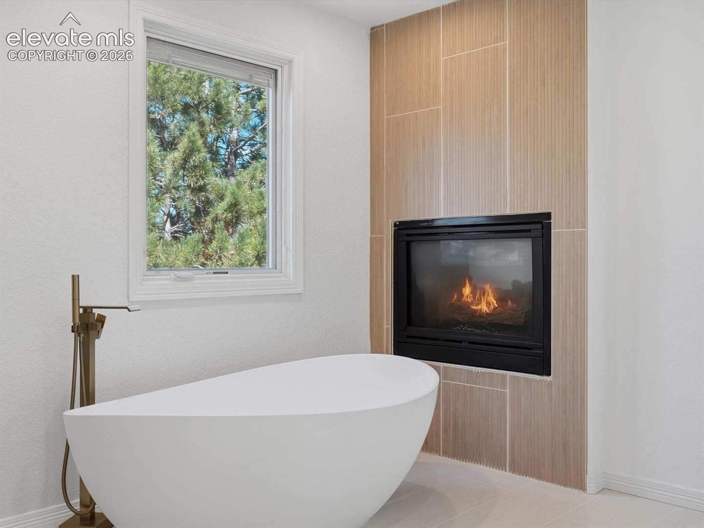 Freestanding soaking tub with a floor-mounted bronze-finish faucet, built-in fireplace featuring vertically installed tile surround, and window with exterior tree-lined backdrop