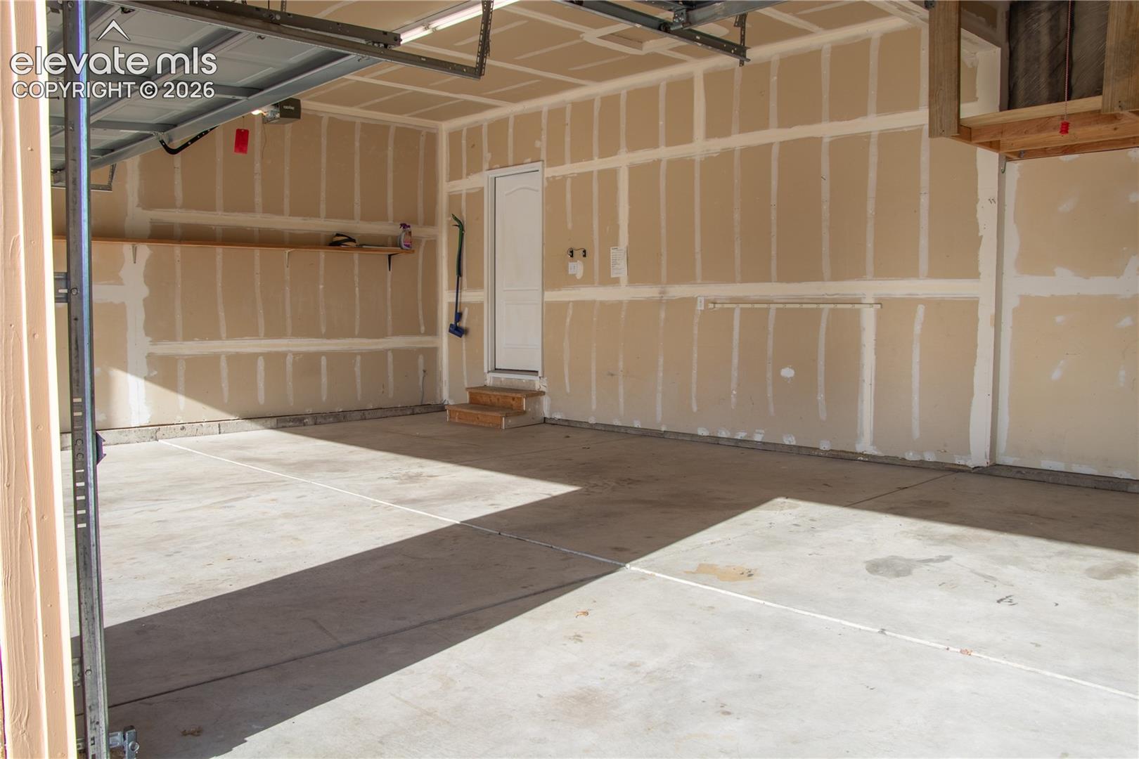 Two-car garage featuring a concrete floor, unpainted drywall with taped seams, an overhead garage door mechanism, and a side entry door with wooden steps