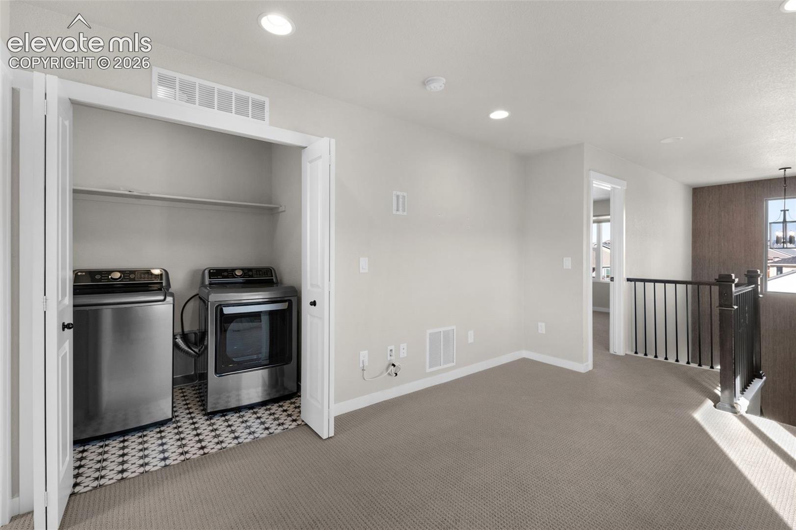 Carpeted landing area featuring a laundry closet with patterned tile flooring, a dark wood newel post and baluster railing, and recessed lighting