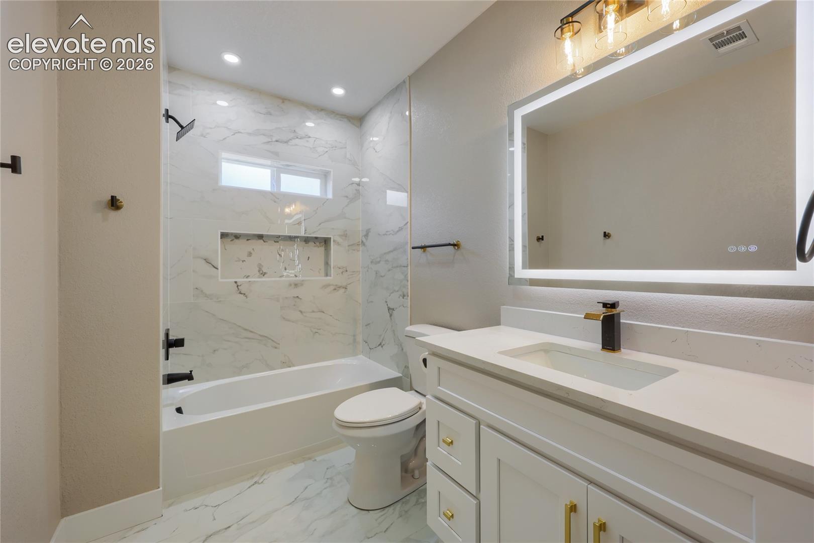 The beautiful and spacious main hall bathroom, features a gorgeous marble tub surround and flooring, quartz countertop, a custom-built shower niche, and contemporary fixtures and finishes.