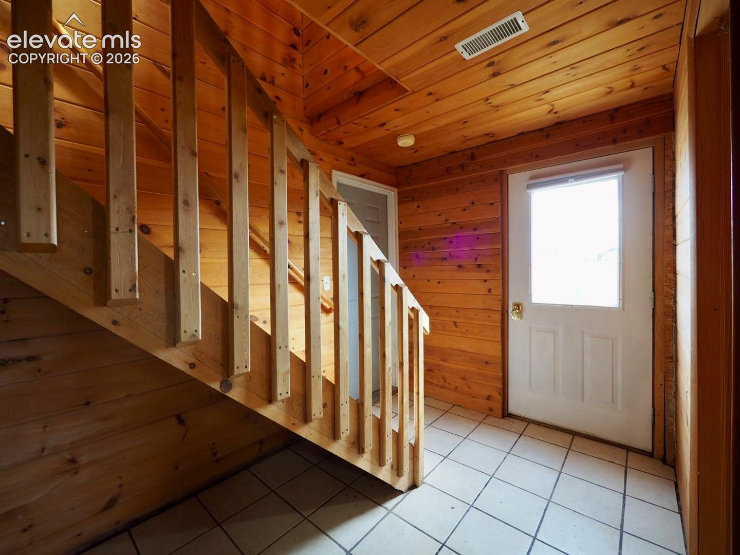 Natural wood paneling on walls and ceilings, staircase with vertical wood balusters, and light-toned ceramic tile flooring