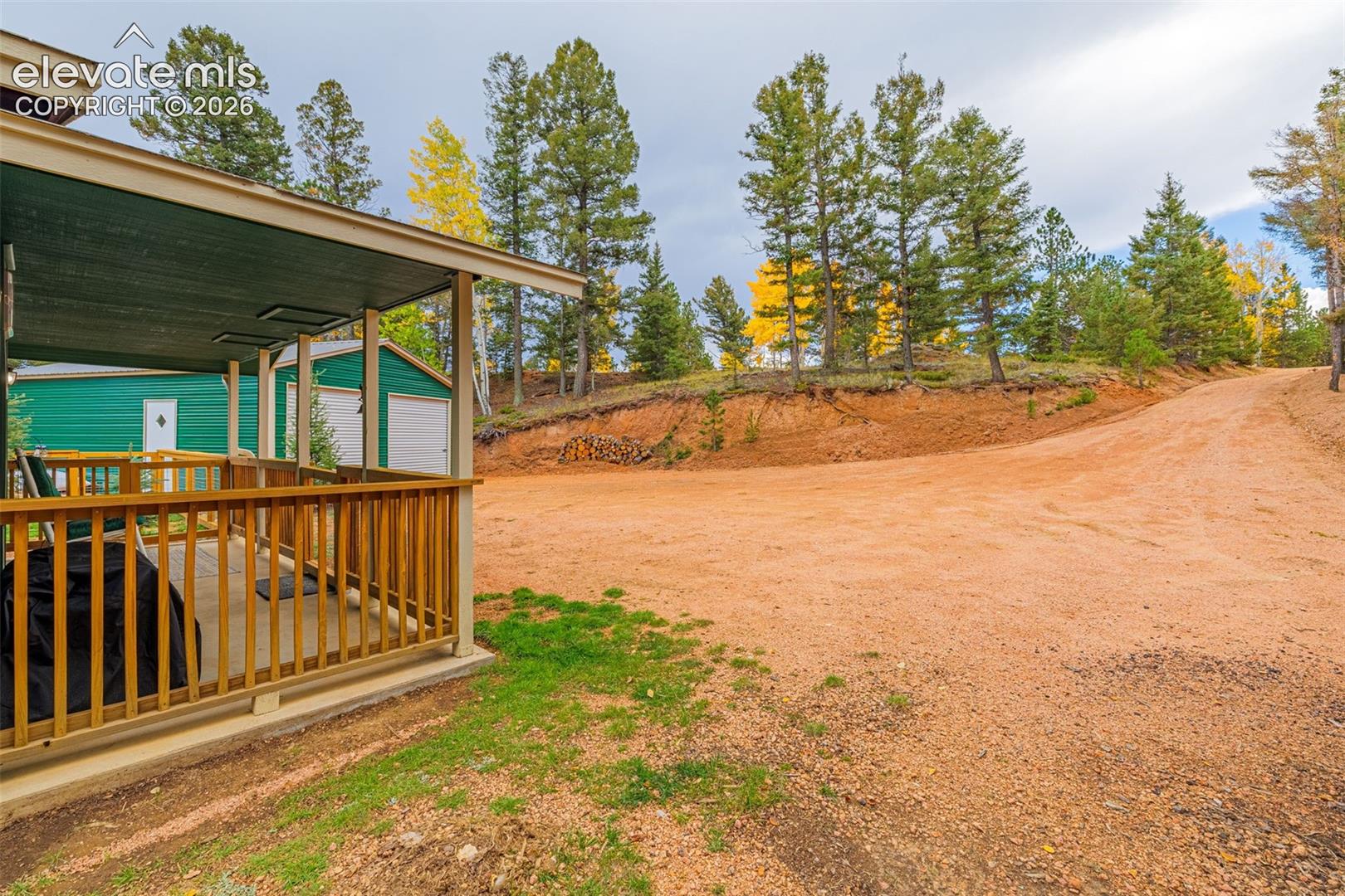 Covered wooden deck featuring vertical balusters, expansive natural grounds, mature pine and aspen trees, and an exterior storage structure with dual garage doors