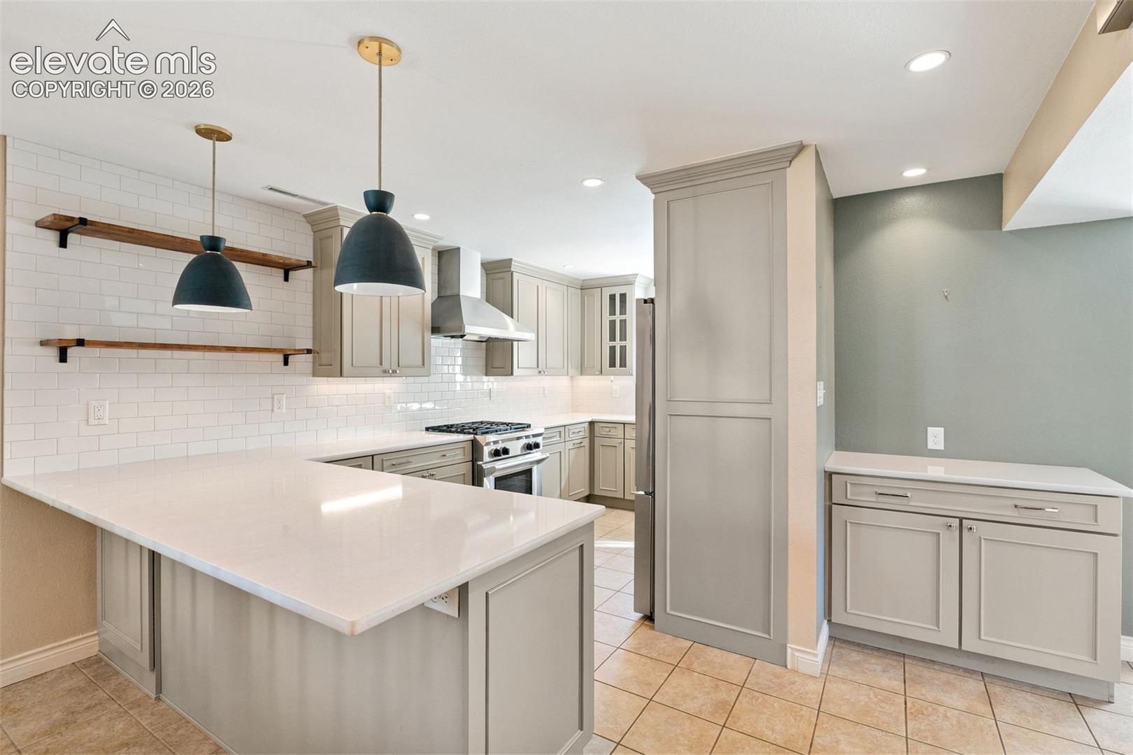 Kitchen featuring white subway tile backsplash, quartz countertops, shaker-style cabinetry, stainless steel range hood, and wood-finish wall shelving