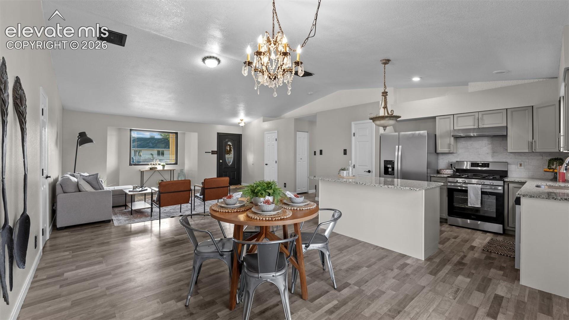 Open-concept living space featuring wood-finish flooring, vaulted ceilings, a kitchen island with stone countertops, stainless steel appliances, and decorative chandeliers
