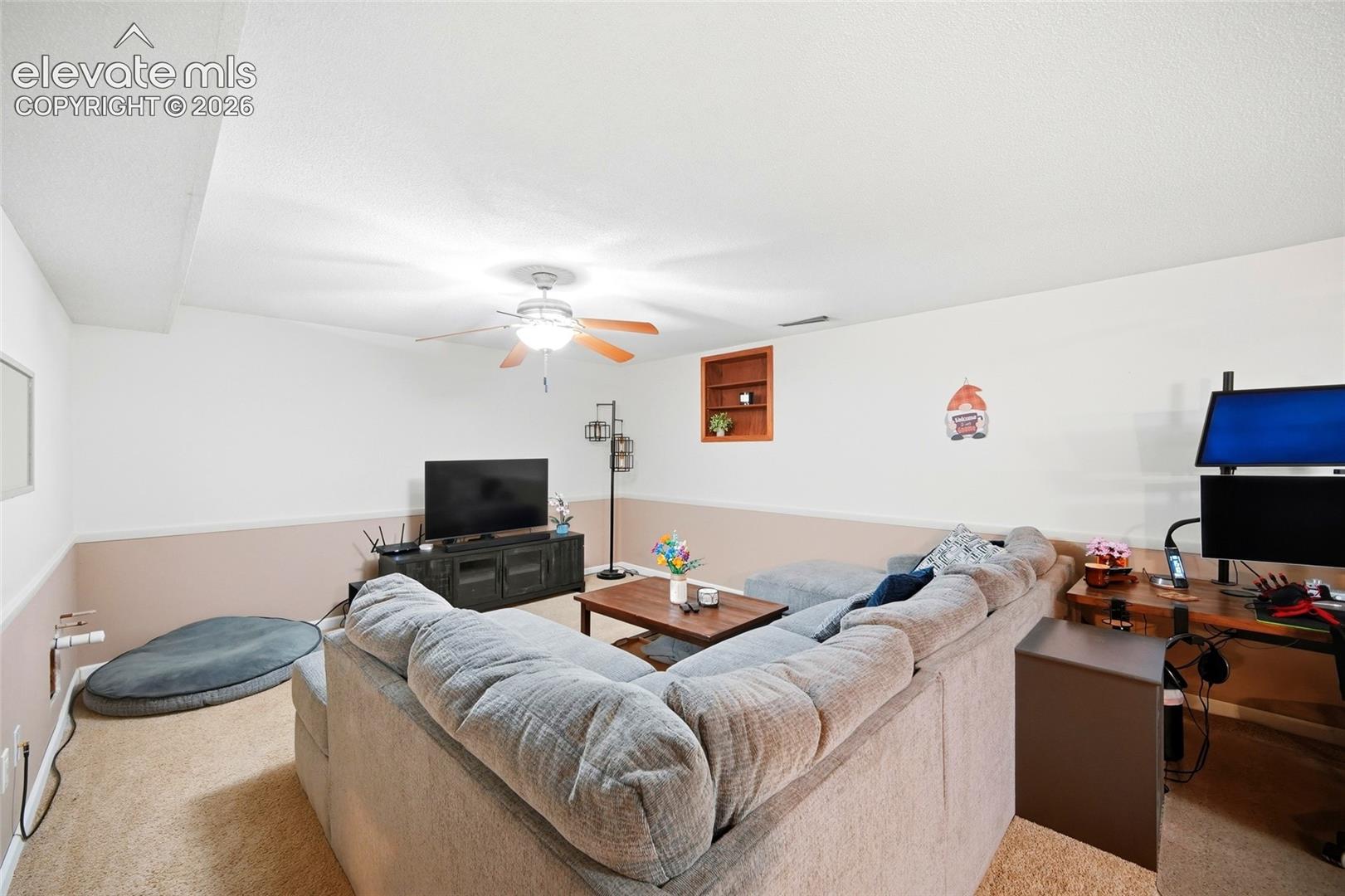 Neutral-toned walls featuring chair rail molding, wall-to-wall carpeting, ceiling fan with light fixture, and recessed wall shelving