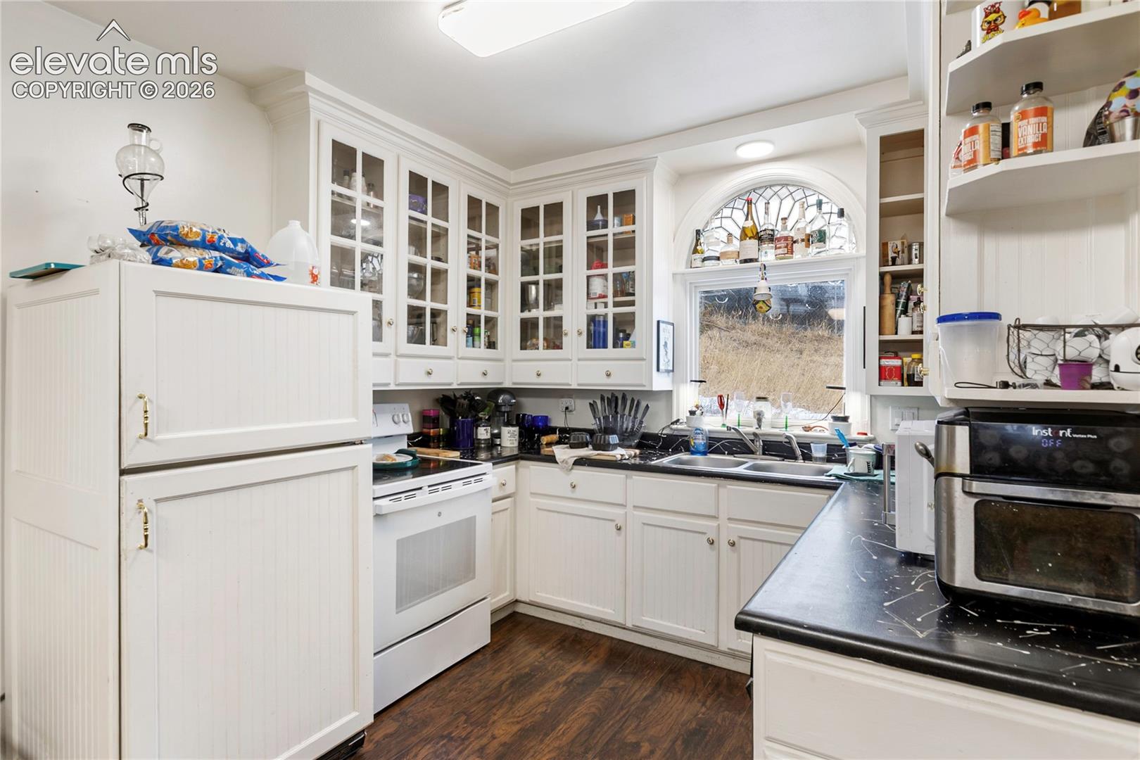 Kitchen featuring white cabinetry with glass-panel upper doors, dark countertops, wood-finish flooring, and an arched window with decorative leaded glass detailing