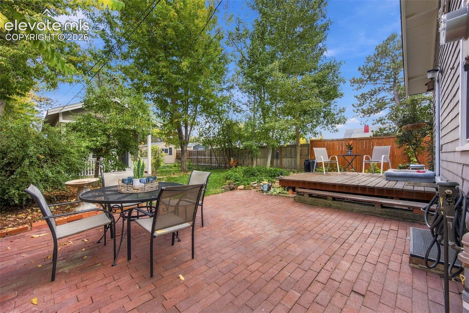Outdoor area featuring a red brick patio, elevated wooden deck, perimeter wood fencing, and mature trees