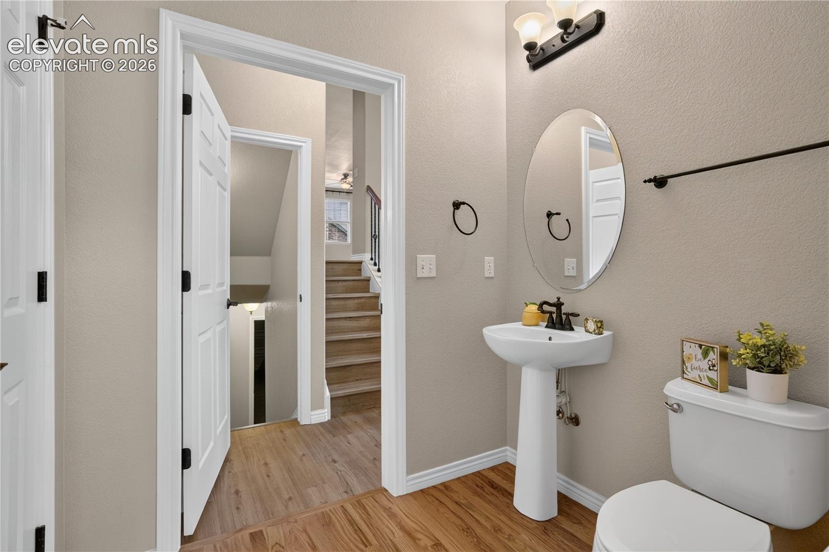 Powder room featuring a white pedestal sink with an oil-rubbed bronze faucet, oval mirror, wall-mounted light fixture, and wood-finish flooring