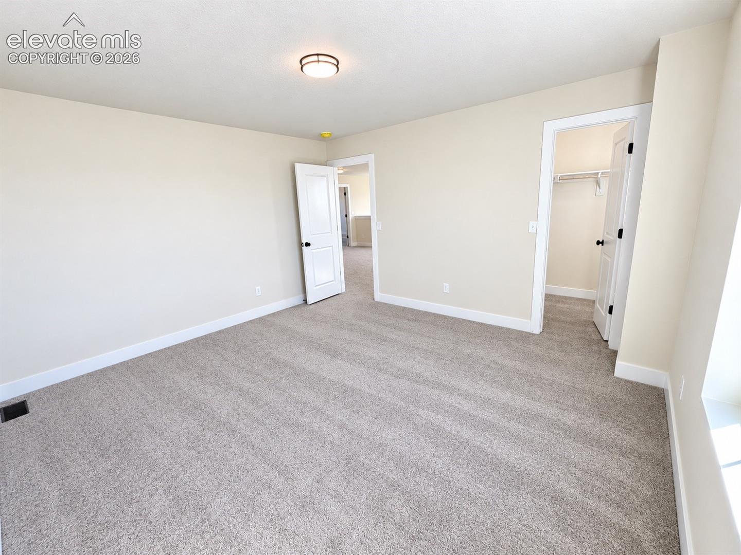 Neutral tone wall paint, light-colored carpeting, white trim, flush mount ceiling light fixture, and a built-in closet with shelving