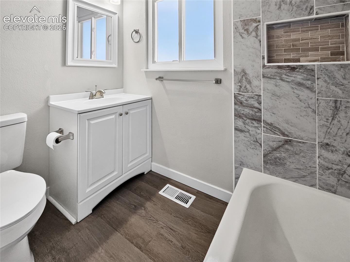 Bathroom featuring a single-sink vanity with white cabinetry, a built-in bathtub, a tiled shower surround with a recessed niche, wood-finish flooring, and a window providing natural light