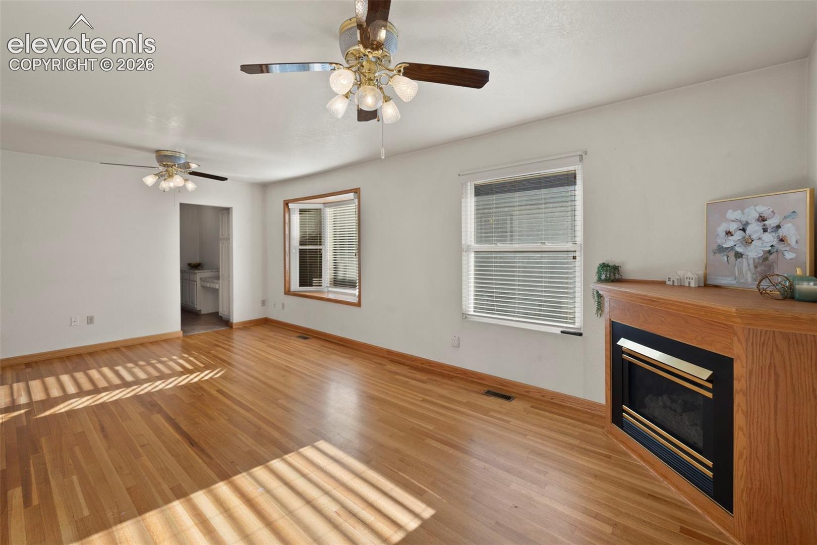 Hardwood flooring throughout, featuring a corner fireplace with a wood mantel, multiple windows with blinds, and ceiling fans with integrated lighting