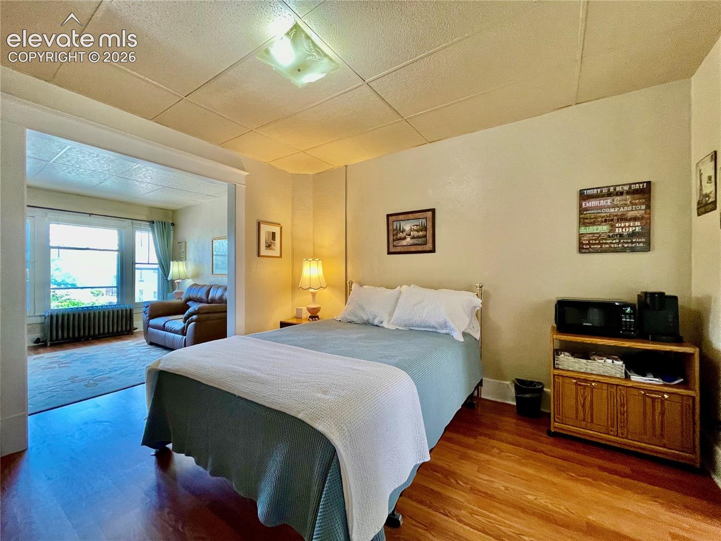 Wood-finish flooring throughout, recessed ceiling light, and an open doorway leading to an additional space with large windows and a radiator