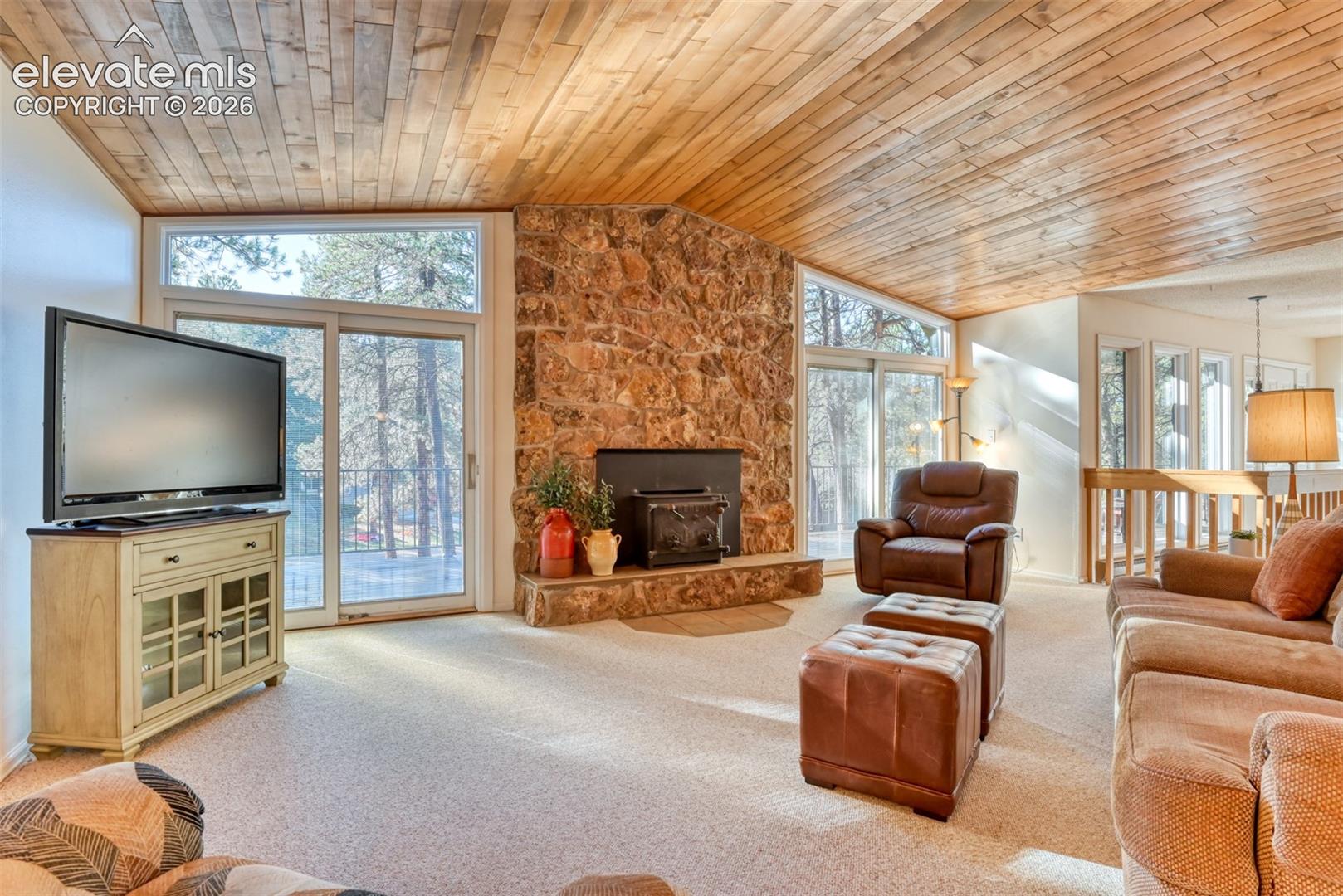 Vaulted wood-paneled ceiling, floor-to-ceiling stone fireplace with a wood-burning stove insert, and expansive window walls with sliding glass doors