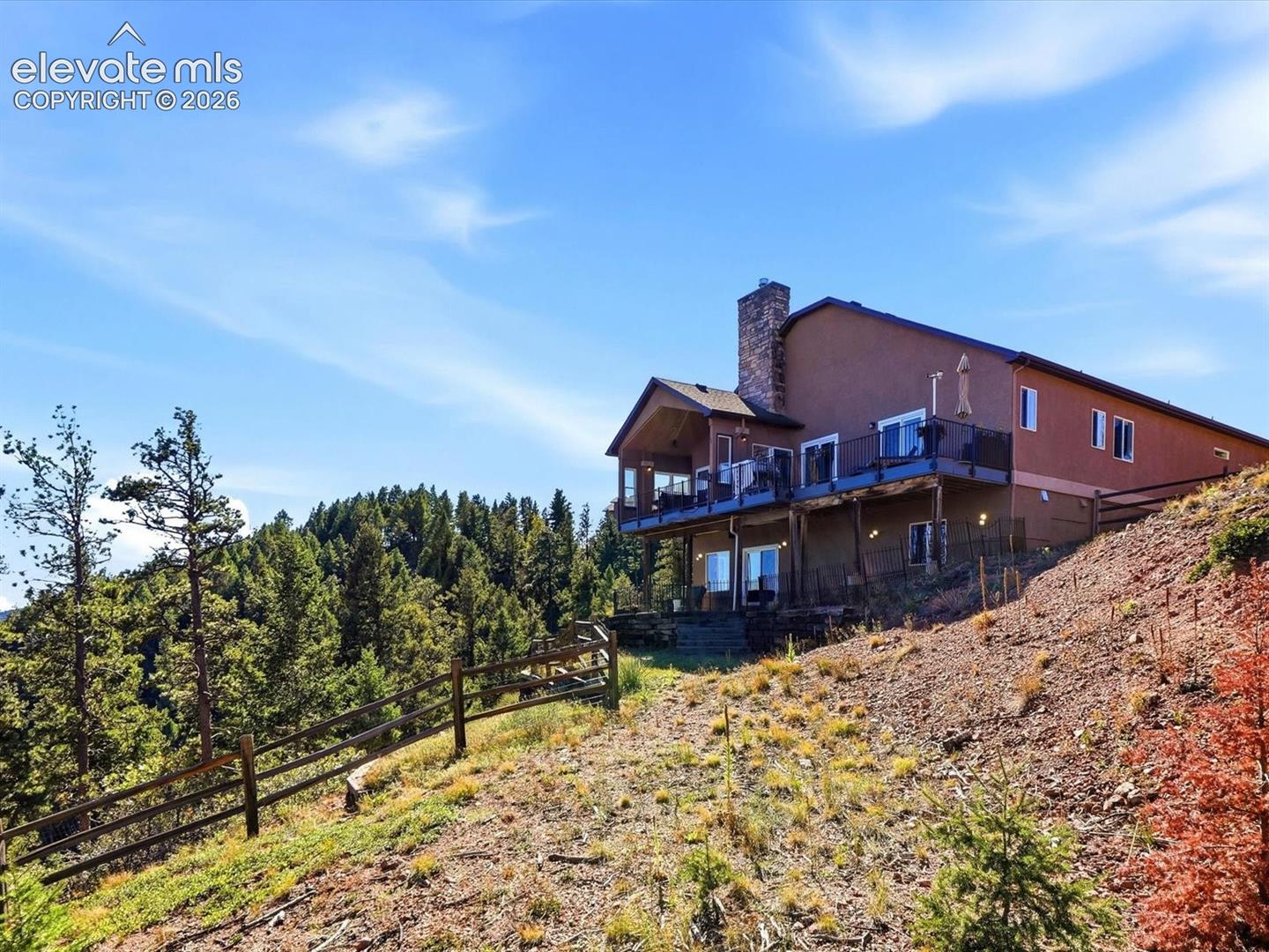 Multi-level exterior featuring a stone-clad chimney, tiered elevated deck with metal railing, and walk-out lower level on a hillside lot with perimeter timber fencing