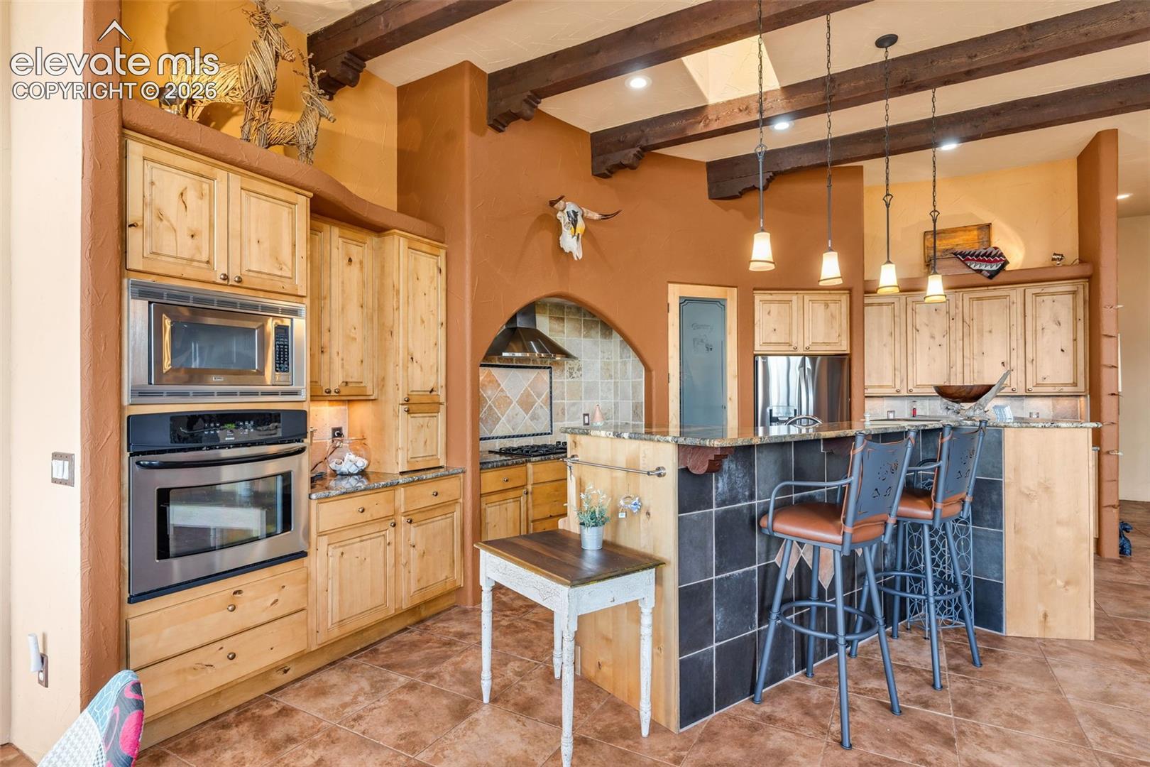 Kitchen featuring light wood cabinetry, stone countertops, stainless steel wall-mounted oven and microwave, dark tile-front breakfast bar, and exposed wood ceiling beams