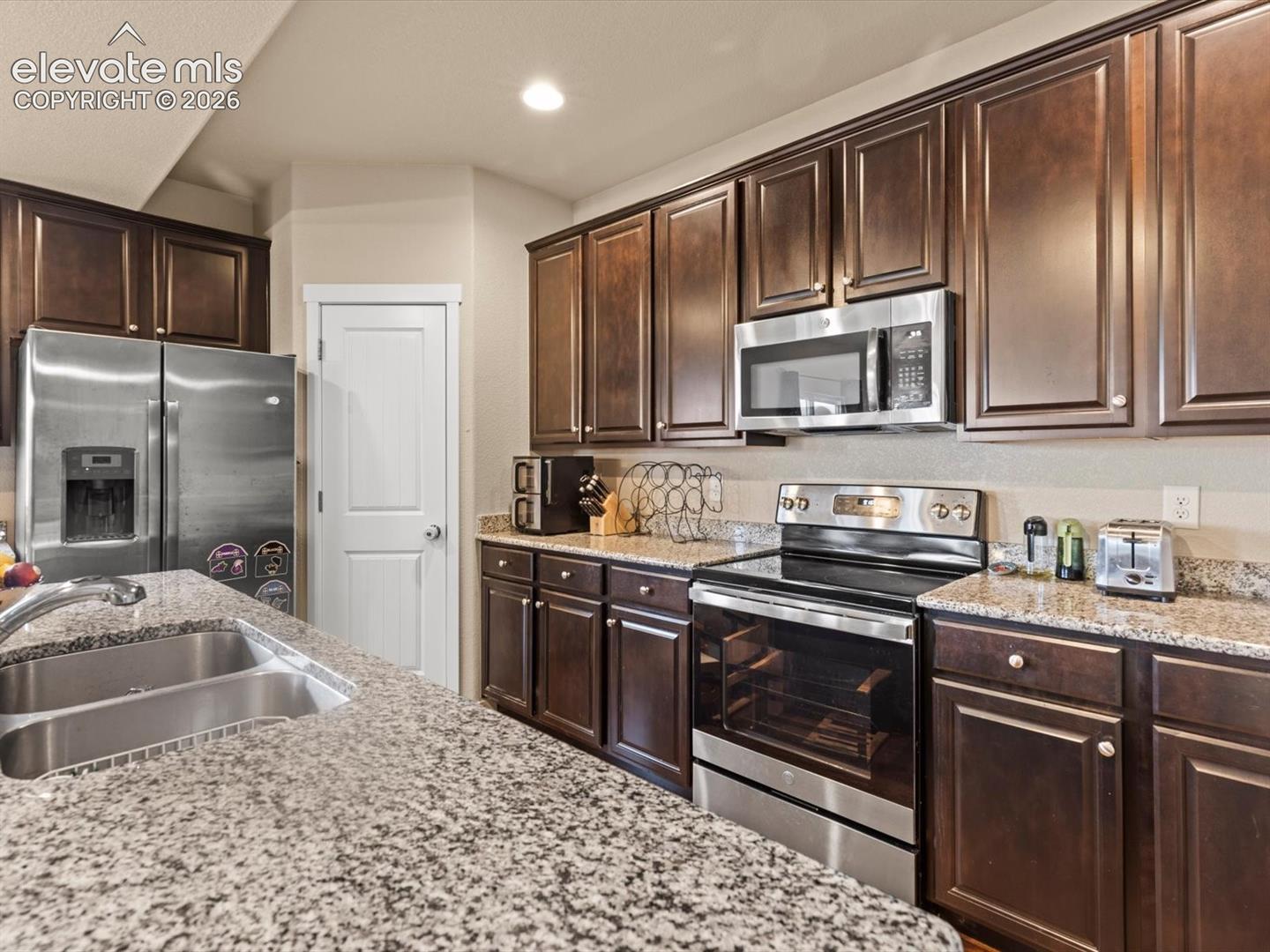 Kitchen featuring extensive granite countertops, dark wood cabinetry, stainless steel appliances, and a double basin sink