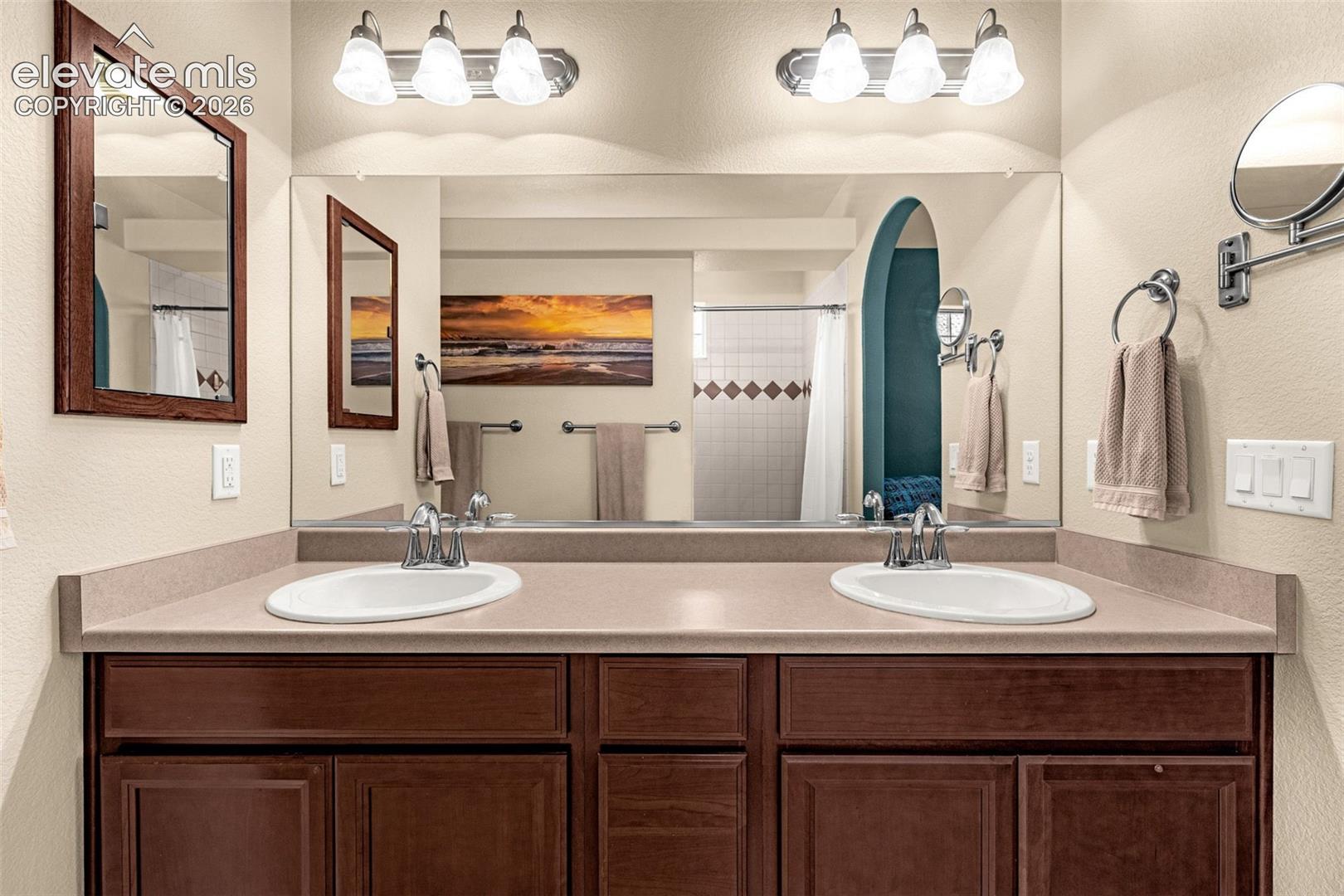 Double-sink vanity featuring a solid surface countertop, wood-finish cabinetry, two dual-light fixtures, and a wall-to-wall mirror