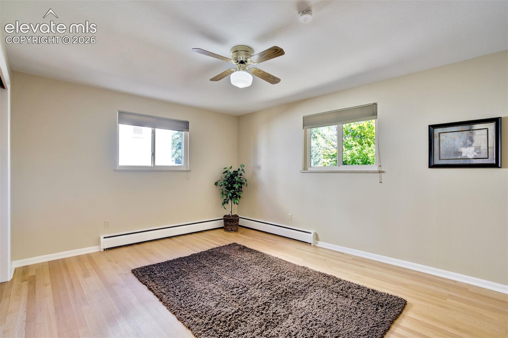 Neutral-toned room featuring wood-finish flooring, dual windows with fitted shades, and a central ceiling fan with integrated lighting