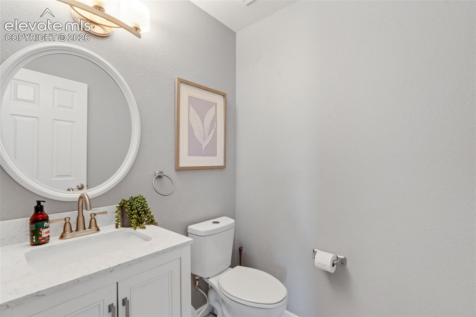 White vanity featuring a stone-look countertop, integrated basin, and warm-toned dual-handle faucet, accented by a circular wall-mounted mirror and two-light fixture